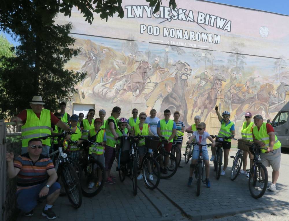 Grupa dzieci, kobiet i mężczyz z rowerami, w tle budynek z muralem Bitwy pod Komarowem